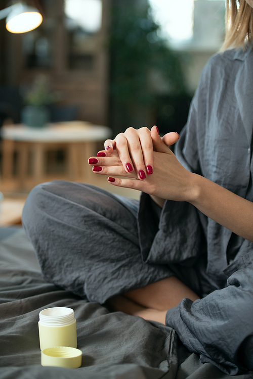 Young female in grey pajamas applying natural handmade cosmetic product on her hands while sitting on bed in the morning