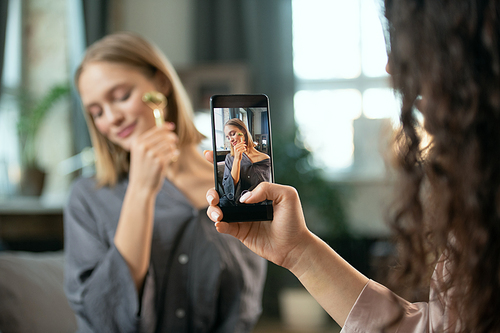 Hand of young woman with smartphone shooting her friend using massage roller upon her face and enjoying beauty procedure