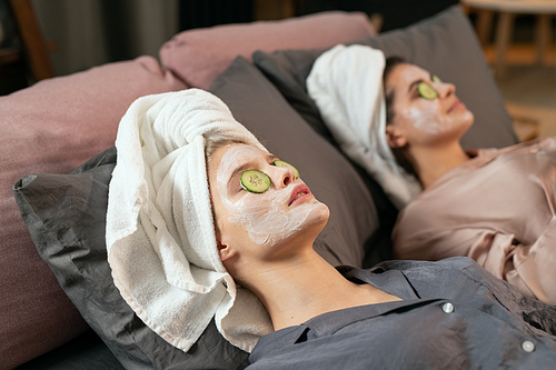 Two girls in silk pajamas, towels on heads, clay masks on faces and cucumber slices on shut eyes lying on bed and enjoying beauty procedure