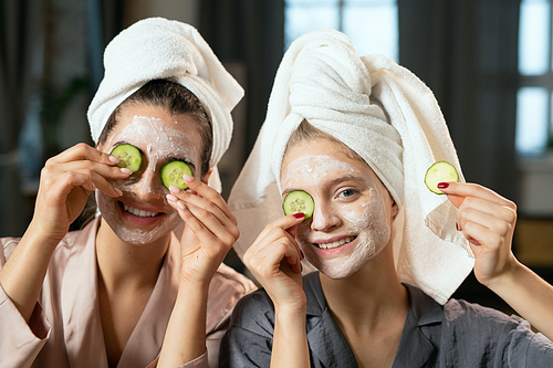 Young women in silk pajamas, towels on heads, clay masks on faces and cucumber slices on eyes having fun and enjoying beauty procedure