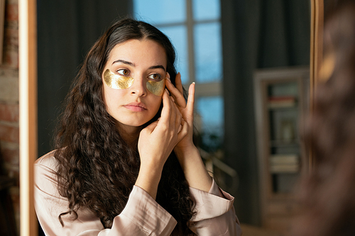 Young brunette woman in silk pajamas putting golden cosmetic undereye patches while standing in front of mirror and looking at herself
