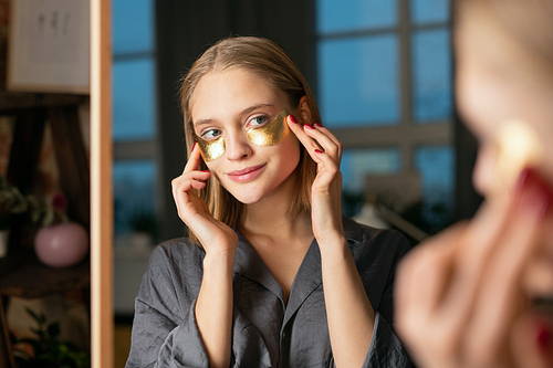 Young smiling blond woman in silk grey pajamas putting golden cosmetic undereye patches while standing in front of mirror and looking at herself