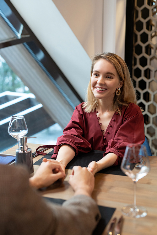 Young blond cheerful female in elegant dress looking at her boyfriend with toothy smile while holding his hands by served table in restaurant