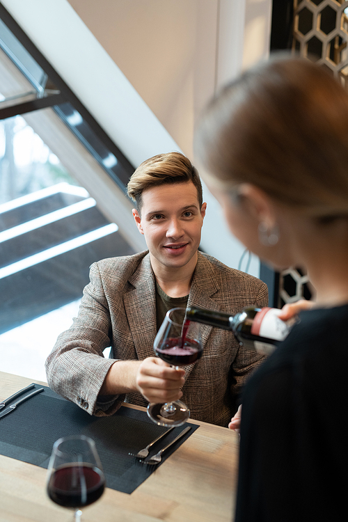 Elegant young man sitting by served table in luxurious restaurant and holding wineglass while waitress pouring red wine into it