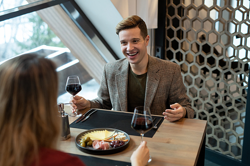 Happy young man with glass of red wine sitting by served table in front of his girlfriend and making toast during romantic dinner in restaurant