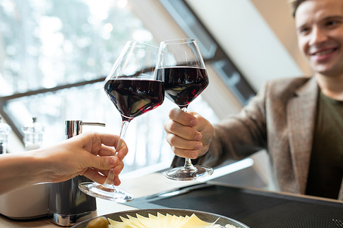 Hands of happy young man and those of his girlfriend clinking by glasses of red wine over served table during romantic dinner in restaurant