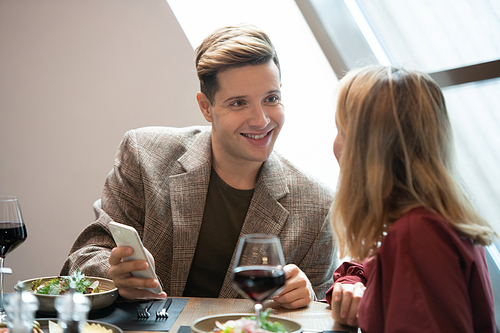 Happy young man with smartphone looking at his girlfriend with smile while talking to her by served table in luxurious restaurant