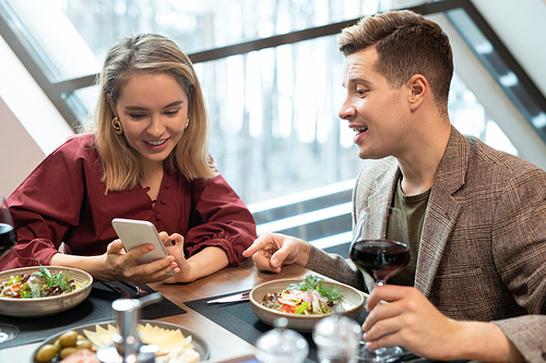 Elegant blond girl with smartphone discussing curious video with her boyfriend while showing it to him by dinner in luxurious restaurant