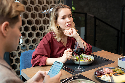 Sulky young blond woman in maroon dress eating salad and expressing sadness because of her boyfriend scrolling in smartphone