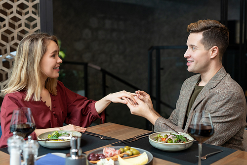 Happy young elegant man looking at his pretty girlfiend with smile while putting engagement ring on her finger over served table by dinner