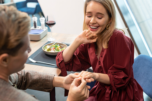 Young cheerful blond woman looking at her finger where her boyfriend putting engagement ring during proposal by dinner in restaurant