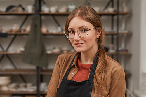 Warm-toned portrait of young female artisan looking at camera while posing in pottery workshop, hobby and small business concept, copy space