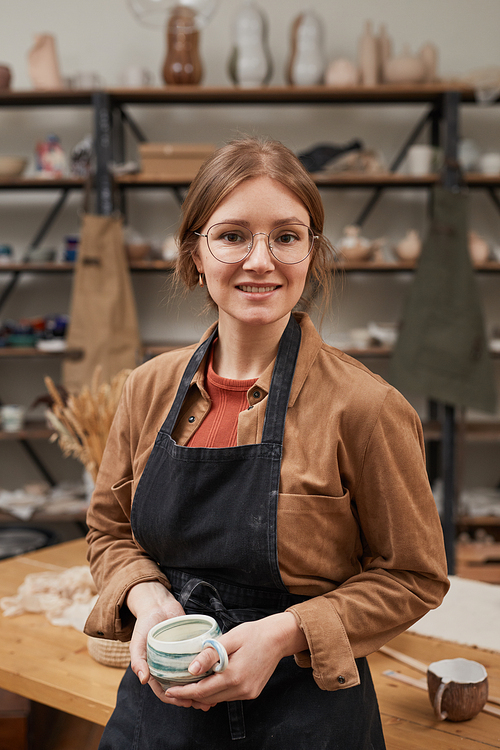 Vertical waist up portrait of young female potter looking at camera while posing in workshop, hobby and small business