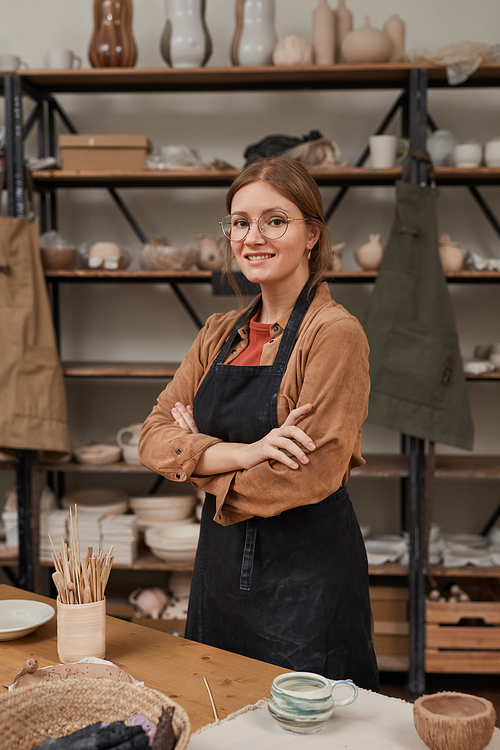 Vertical waist up portrait of young female artisan looking at camera while standing with arms crossed in workshop, hobby and small business concept