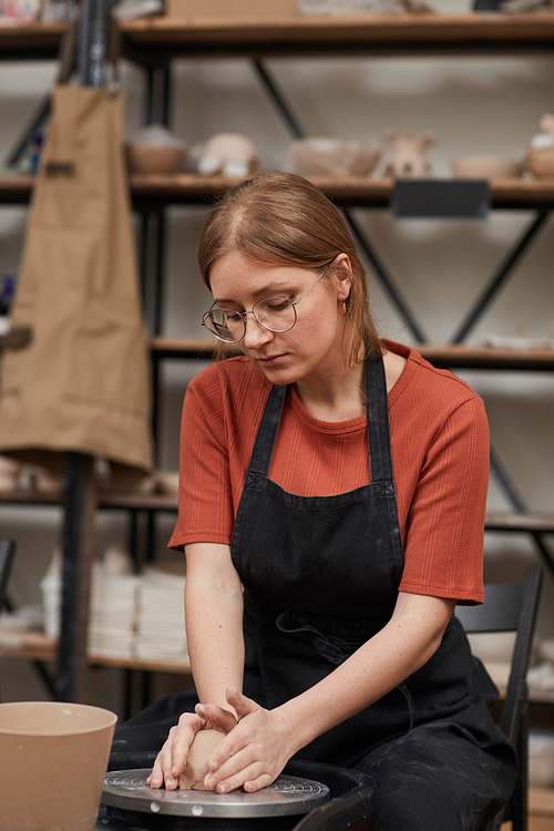 Vertical warm-toned portrait of young woman shaping clay on pottery wheel while enjoying hobby in workshop