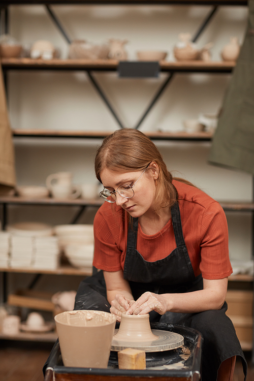 Vertical warm toned portrait of young woman shaping clay on pottery wheel in workshop while enjoying arts and crafts, copy space