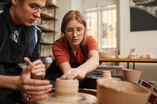 Portrait of female potter helping young man shaping clay on pottery wheel in workshop, copy space