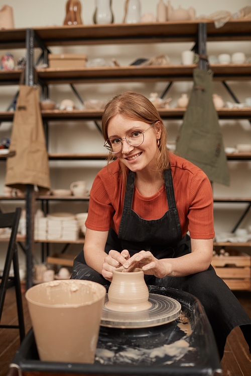 Vertical warm toned portrait of young female potter smiling at camera while working on pottery wheel in workshop and enjoying arts and crafts, copy space
