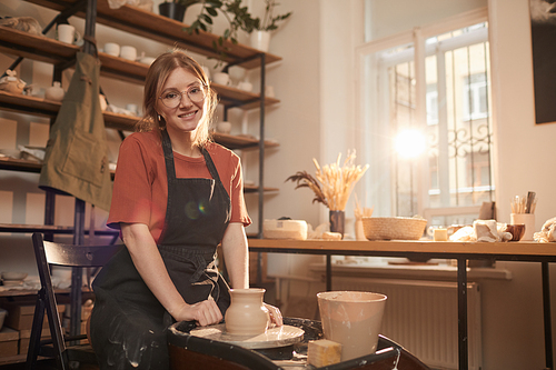 Warm toned portrait of young female artisan smiling at camera while working on pottery wheel in sunlit workshop and enjoying arts and crafts, copy space