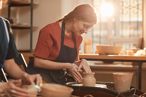 Warm toned portrait of young woman shaping clay on pottery wheel in sunlit workshop and enjoying arts and crafts, copy space