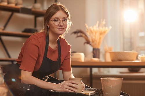 Warm toned portrait of young woman shaping clay on pottery wheel while making ceramics in sunlit workshop, copy space