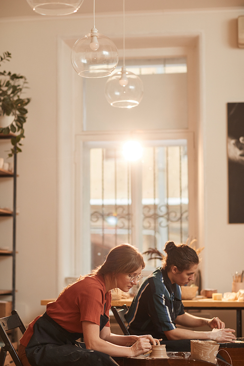 Vertical side view portrait of two young people shaping clay on pottery wheel while making ceramics in workshop lit by sunlight, copy space above