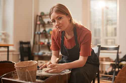 Warm-toned portrait of young female artisan shaping clay on pottery wheel and looking at camera while making ceramics in workshop lit by sunlight, copy space