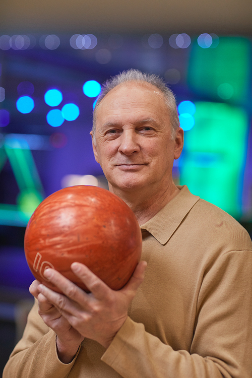 Vertical waist up portrait of smiling senior man holding bowling ball and looking at camera while enjoying active entertainment at bowling alley