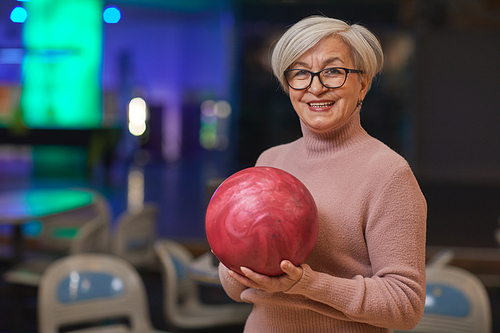 Waist up portrait of smiling senior woman holding bowling ball and looking at camera while enjoying active entertainment at bowling alley, copy space