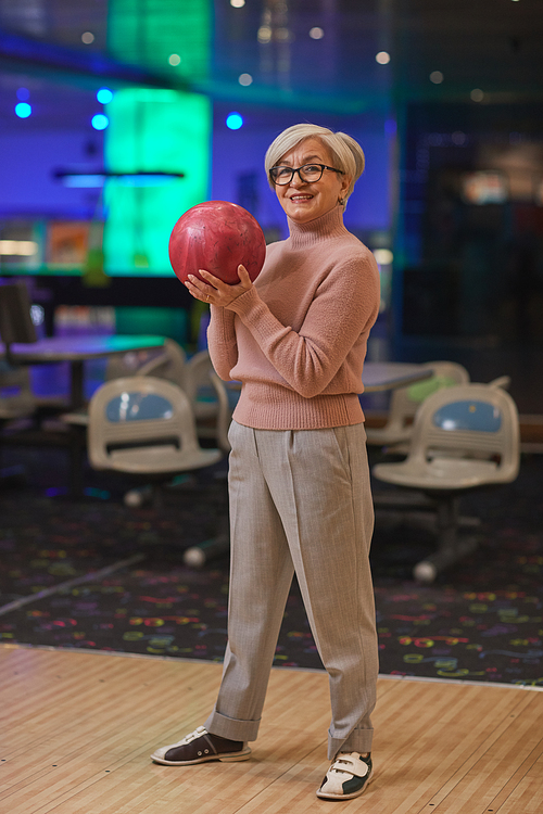 Vertical full length portrait of smiling senior woman holding bowling ball and looking at camera while enjoying active entertainment at bowling alley