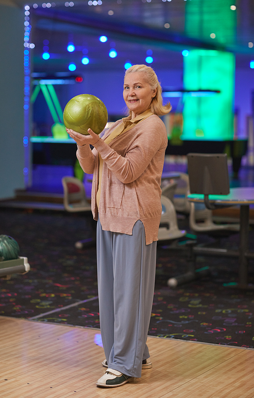 Vertical full length portrait of elegant senior woman holding bowling ball and smiling at camera while enjoying active entertainment at bowling alley