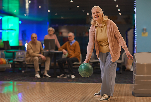 Full length portrait of elegant senior woman playing bowling with group of friends in background, copy space