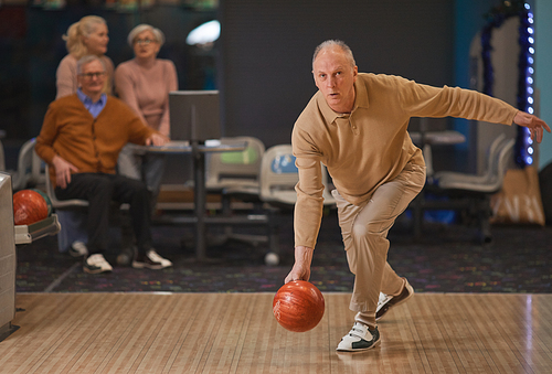 Full length portrait of active senior man playing bowling and throwing ball by lane with group of friends in background, copy space