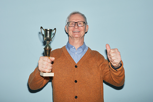 Waist up portrait of smiling senior man holding trophy and looking at camera while standing against blue background, copy space