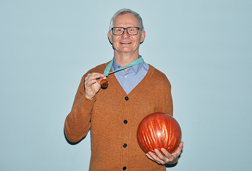 Waist up portrait of smiling senior man holding bowling ball and wearing medal smiling at camera while standing against blue background, copy space