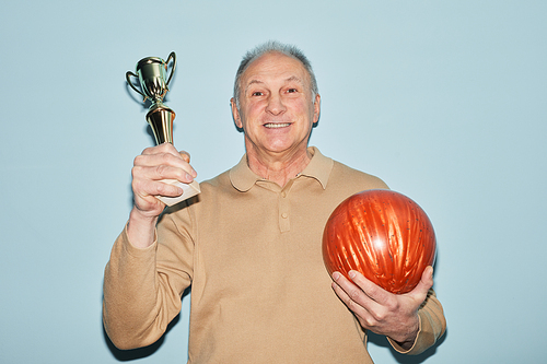 Waist up portrait of smiling senior man holding trophy and bowling ball while standing against blue background, shot with flash, copy space