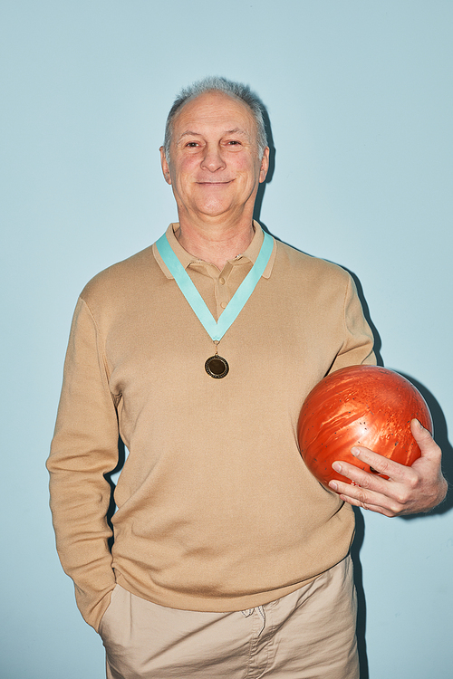 Vertical waist up portrait of smiling senior man holding bowling ball while standing against blue background, shot with flash