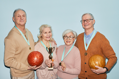 Waist up portrait of group of senior people holding trophy and bowling balls while standing against blue background, shot with flash