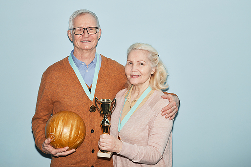 Waist up portrait of smiling senior couple holding trophy and bowling balls while standing against blue background, shot with flash, copy space