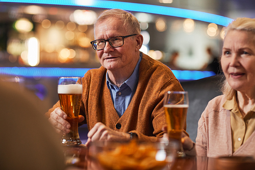 Portrait of smiling senior man drinking beer in bar while enjoying night out with friends, copy space