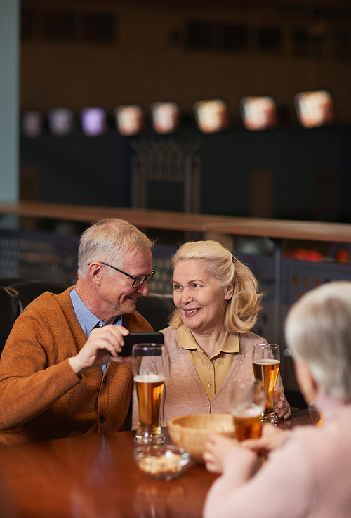 Vertical portrait of modern senior couple taking selfie photo while drinking beer in bar and enjoying night out with friends, copy space