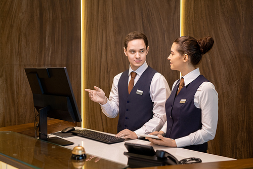 One of two young hotel receptionists standing by counter, looking at colleague with touchpad and pointing at computer screen