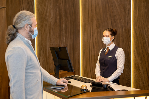 Young female receptionist in uniform and protective mask standing by counter and looking at mature businessman while consulting him