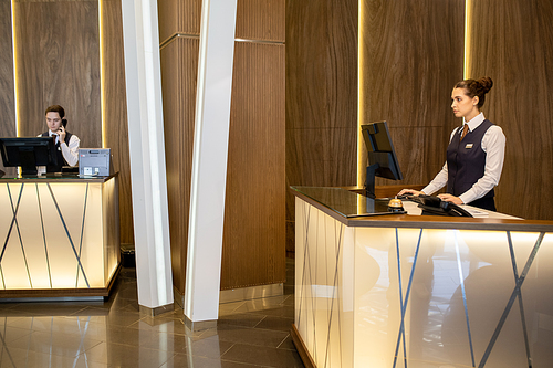 Young elegant female receptionist in uniform standing by counter and looking at computer screen while her colleague answering phone