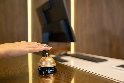 Hand of young woman over bell on wooden reception counter going to push button and call for receptionist to registrate her in hotel room