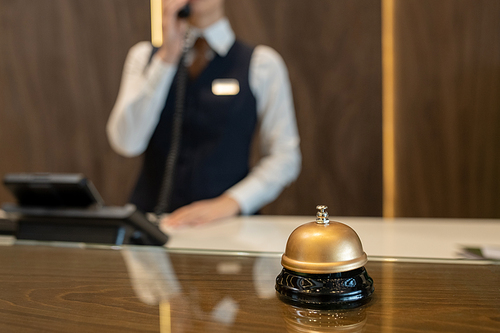 Bell on wooden reception counter against female receptionist consulting clients on the phone while working in contemporary luxurious hotel