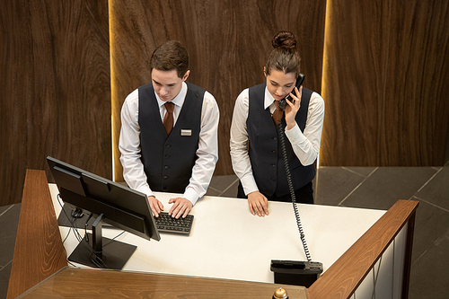 One of two young hotel receptionists standing by counter and looking at computer screen while his colleague answering phone near by