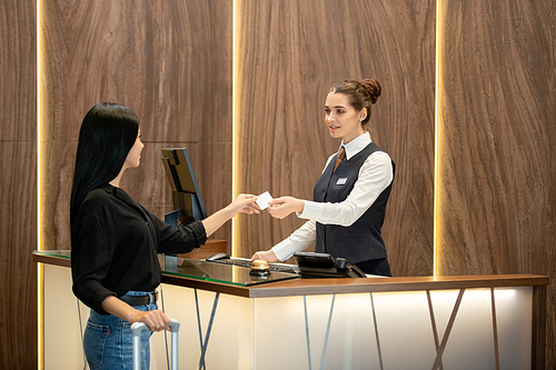 Young brunette receptionist in uniform standing by workplace and passing plastic card to one of guests over reception counter in hotel lobby