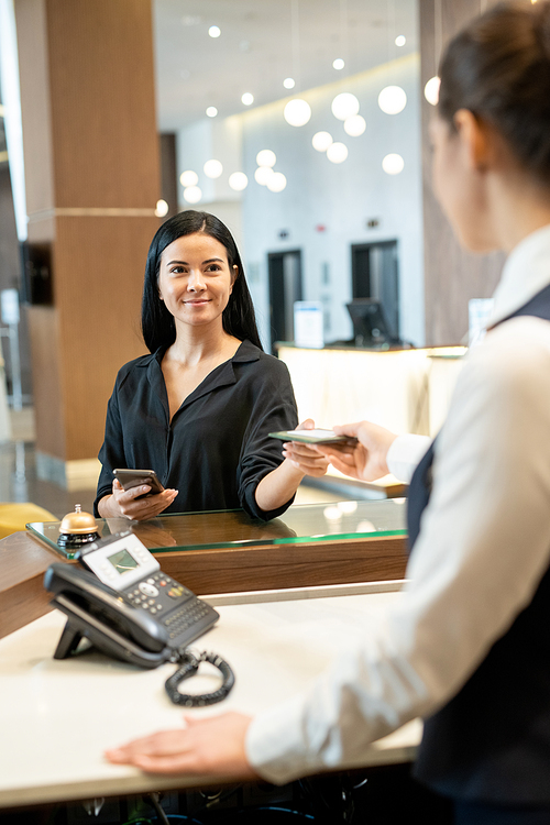 Young brunette businesswoman in casualwear taking documents from hand of female receptionist standing by counter in hotel lobby