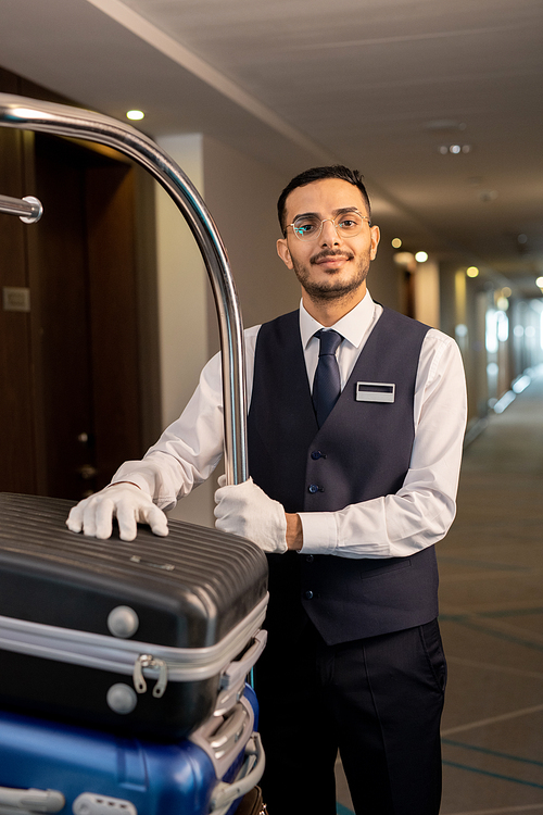 Young serious porter in uniform and eyeglasses standing by cart with suitcases of guests in front of camera in corridor of contemporary hotel